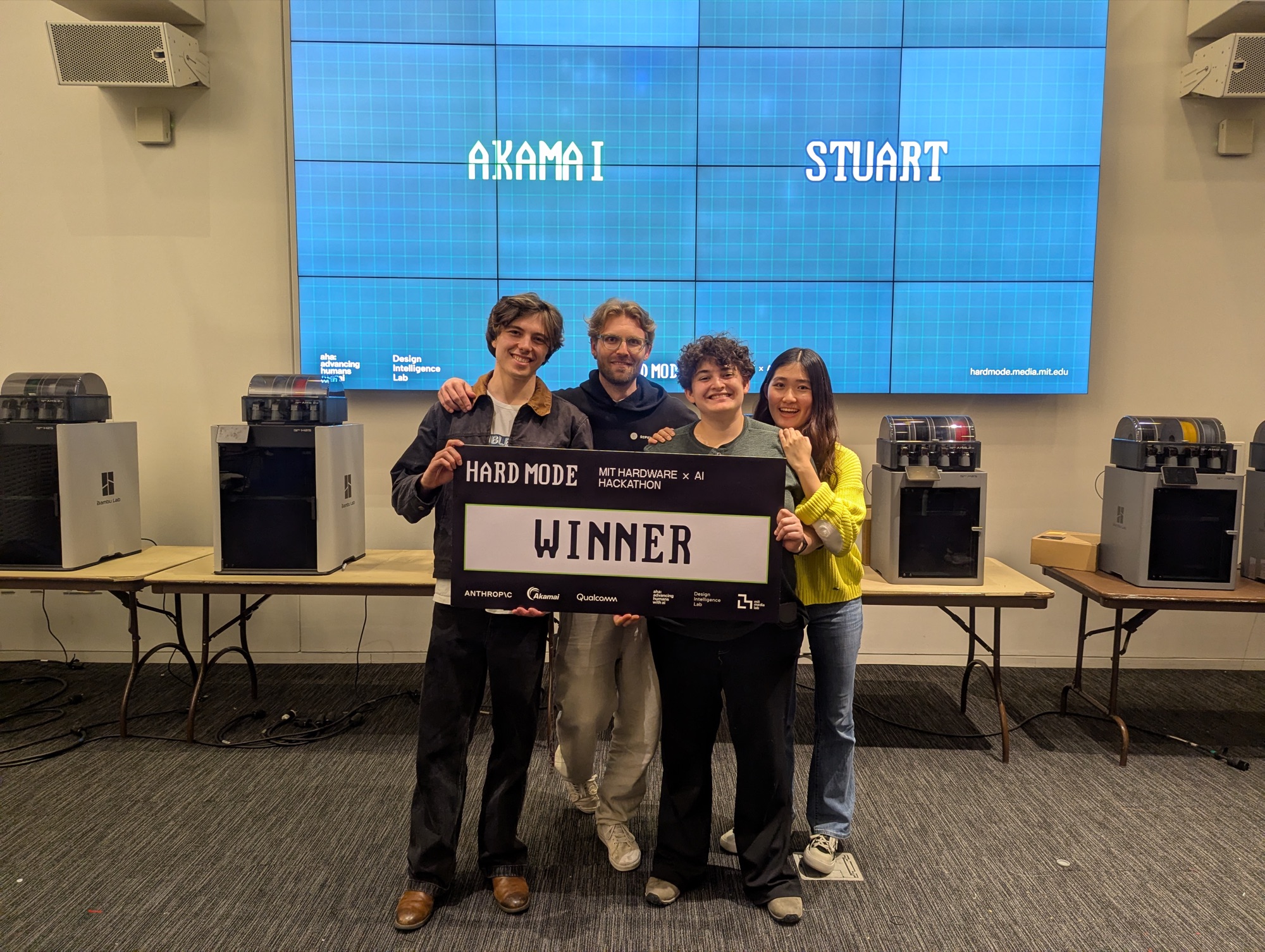The Stuart team holding a HARD MODE 'Winner' sign at the MIT Hardware Hackathon, with AKAMAI and STUART on the screens behind them